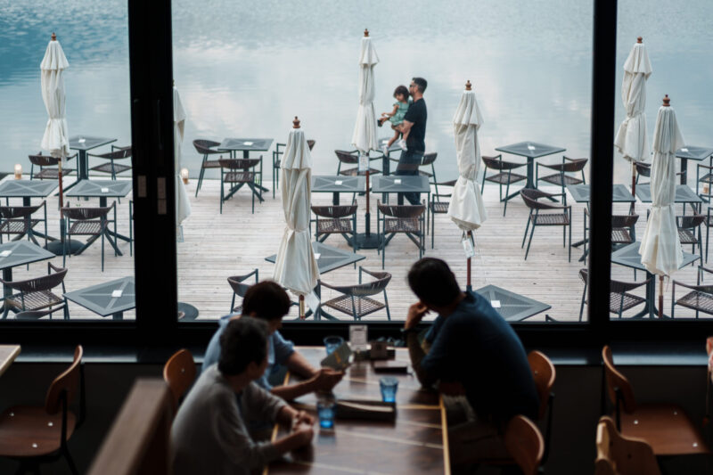 People sitting inside Ao Lakeside Café overlooking outdoor terrace seating and Lake Aoki in Hakuba, Japan