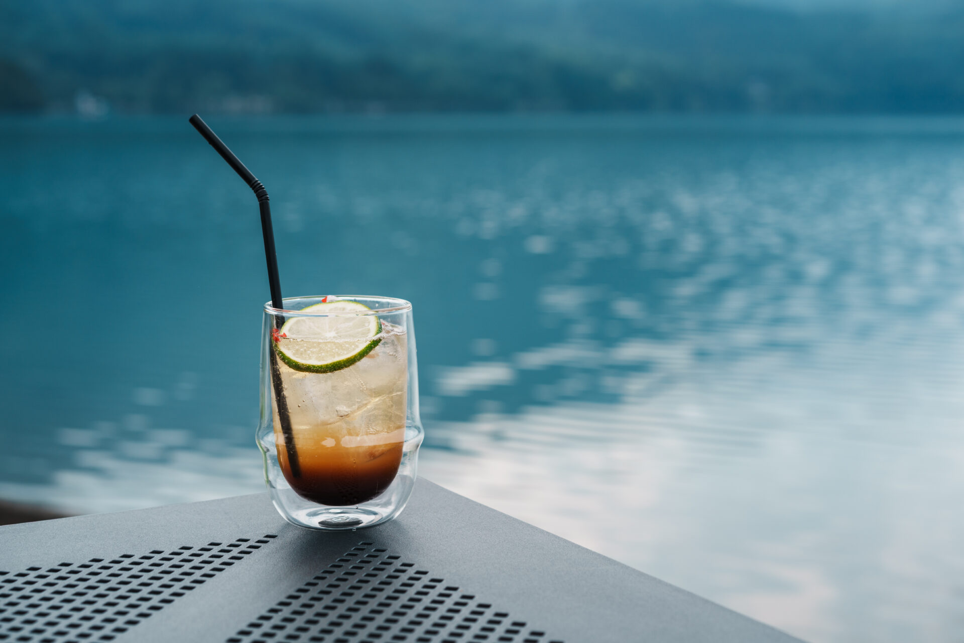 Iced cocktail with lime slice and black straw on a lakeside terrace table overlooking Lake Aoki in Hakuba, Japan