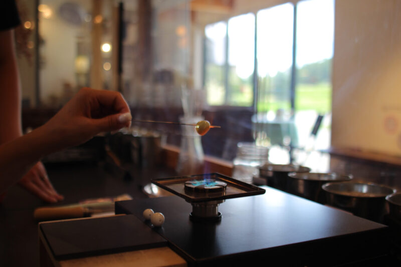 Glass bead being melted over a flame in the Kira Kira Glass workshop, backlit by afternoon sunlight coming in from the floor to ceiling windows in the studio.