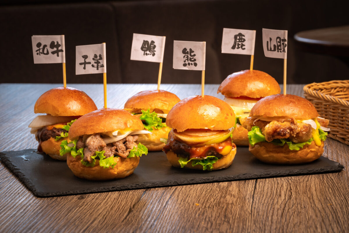 Six wild game sliders from Bears Café Kumaza in Hakuba, Japan, featuring wagyu, lamb, duck, bear, venison, and sanzoku meat, each labeled with handwritten Japanese flags.