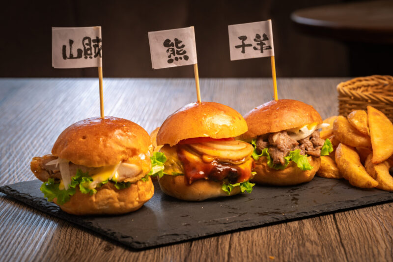 Trio of wild meat sliders from Bears Café Kumaza in Hakuba, Japan, featuring sanzoku-style chicken, bear burger with grilled onion and sauce, and lamb, each labeled with handwritten Japanese flags.