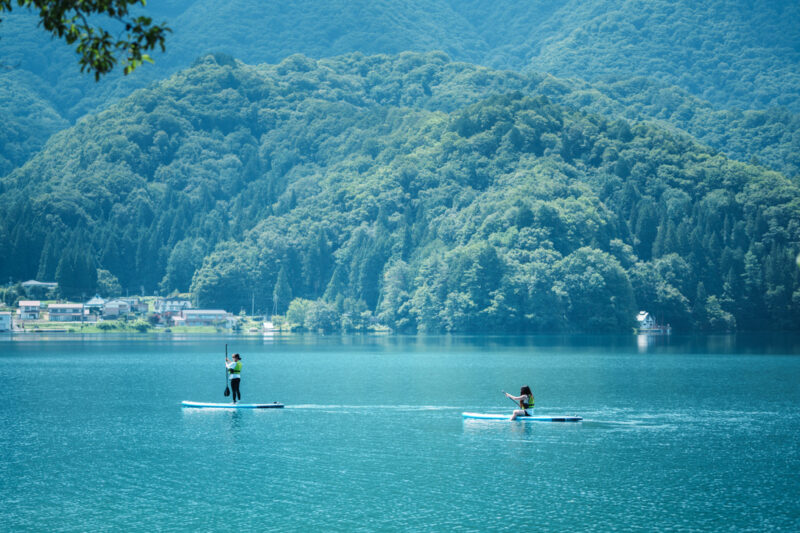 Two people enjoying stand-up paddleboarding on the calm turquoise waters of Lake Aoki in Hakuba, Nagano, Japan, surrounded by lush green mountains on a summer day.