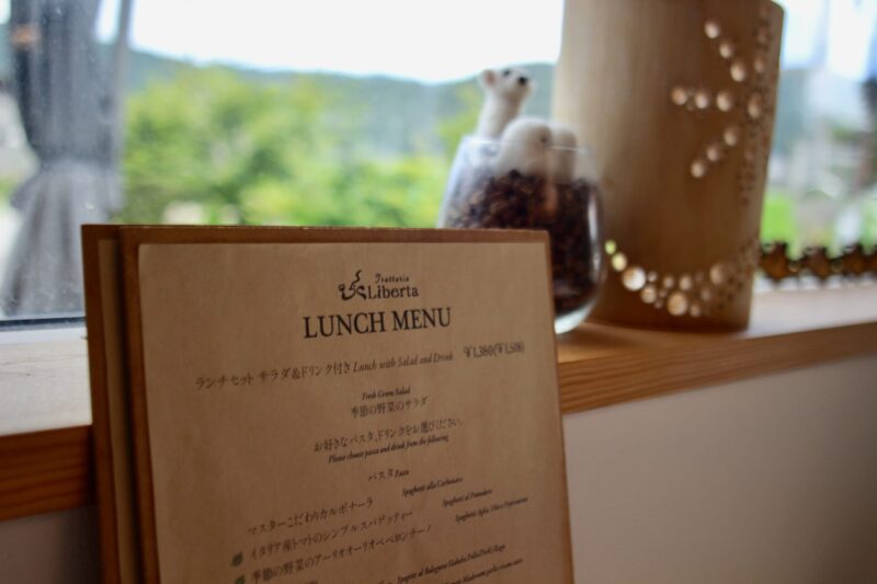 Bulletin board at Gravity Worx Hakuba showcasing colorful food photos, handwritten menu items, and playful illustrations — highlighting the café’s homemade pasta dishes, seasonal specials, and cozy mountain vibe with jars of aged chili oil displayed nearby. 