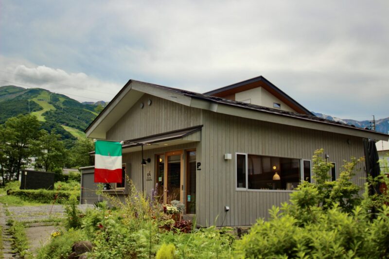Entrance to Gravity Worx Hakuba, a rustic log cabin restaurant surrounded by seasonal alpine flowers and lush greenery near Hakuba Station.