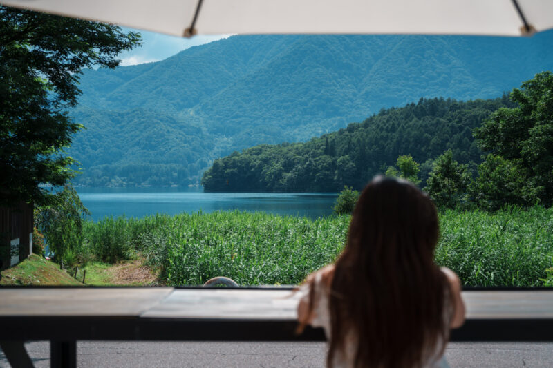 A visitor enjoys a peaceful view of Lake Aoki in Hakuba, Nagano, Japan, framed by mountains and greenery on a clear summer day.