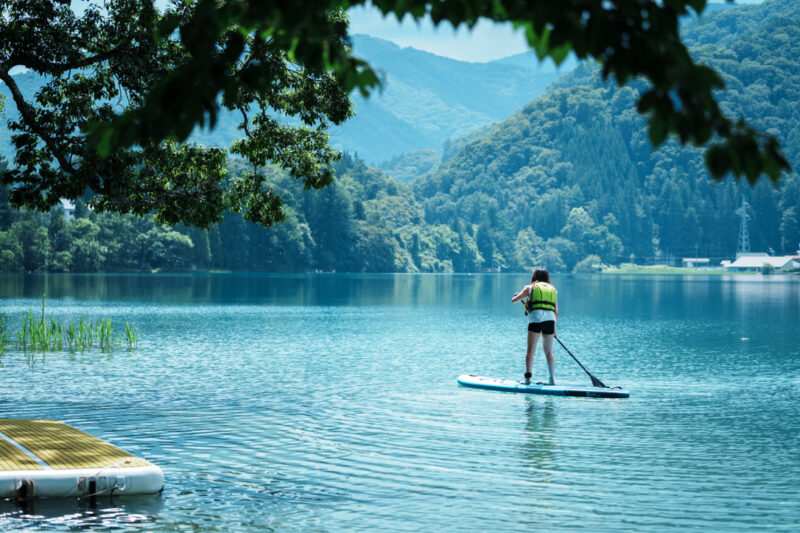 A woman stand-up paddleboarding on the calm blue waters of Lake Aoki in Hakuba, surrounded by lush summer mountains in Nagano, Japan.