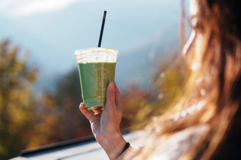 Hand holding a CHAVATY HAKUBA iced matcha latte with mountain views in the background — premium tea latte specialty café in the Japanese Alps, offering scenic outdoor experiences and artisanal drinks made with carefully selected tea leaves.